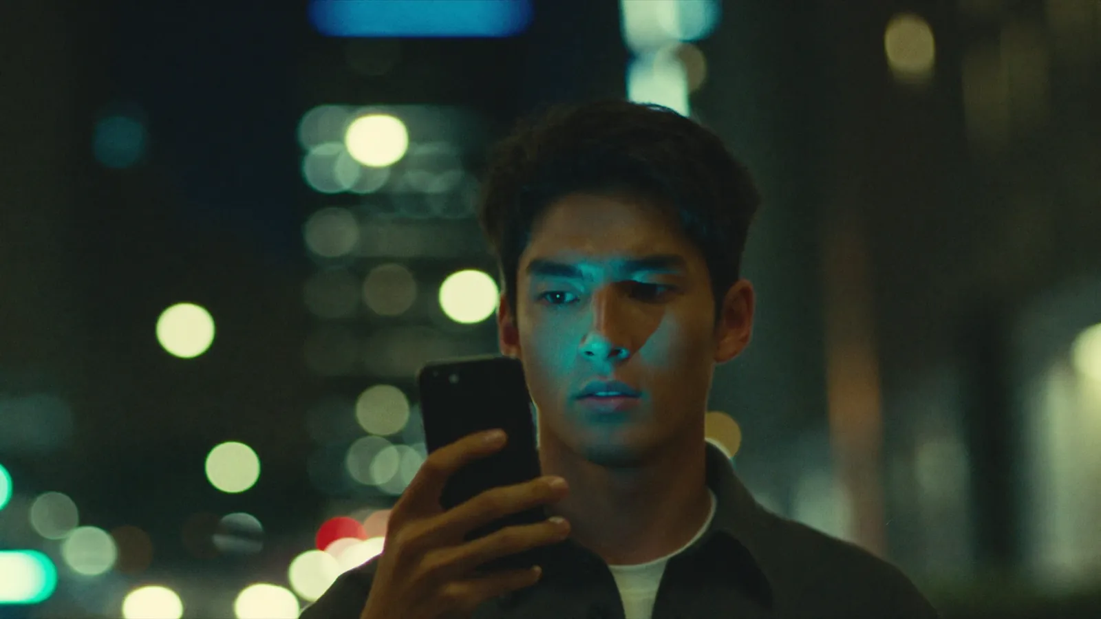 A man on a city street at night asking his phone a question, its light on his face
