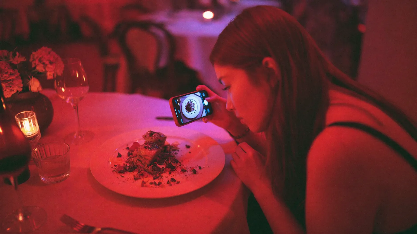 A woman photographing a plated dish with her phone at a candlelit restaurant table