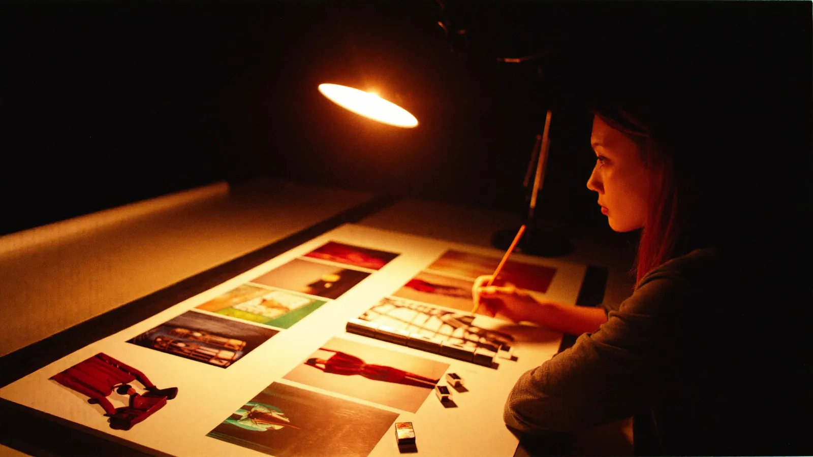A designer arranging printed photographs into a layout under a desk lamp, late at night